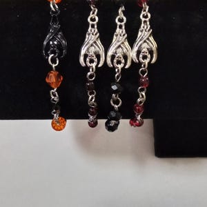 May include: Four pairs of dangle earrings with a Halloween theme. One pair is black with orange beads, and the other three pairs have silver bat charms with black and red beads. The earrings are displayed on a black surface.