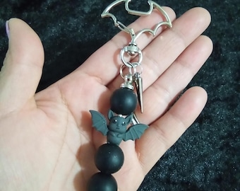 May include: A silver bat-shaped keychain with a black beaded charm. The charm features a small bat figure with outstretched wings and three black beads. A silver spike hangs from the bottom.