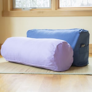 May include: Two yoga bolsters, one lavender and one blue, are lying on a woven mat. The blue bolster has a zipper and a small pocket with a logo.