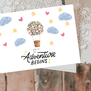 May include: A white card with a hot air balloon illustration, surrounded by clouds, stars, and hearts. The text "...and so the adventure begins" is written in black cursive.