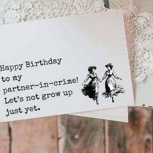May include: A birthday card with a black and white illustration of two women dancing. The card reads "Happy Birthday to my partner-in-crime! Let's not grow up just yet."