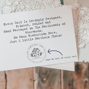 May include: A white card with black text that reads "Every Card Is Lovingly Designed, Printed, Folded and Hand Packaged in The Northwoods of Wisconsin. No Mass Production Here, Just A Little Northern Charm!"  A black and white logo of a house with the text "The Homestead Card Co." is in the lower right corner of the card.