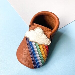May include: A brown leather baby moccasin with a rainbow and cloud design. The moccasin has a white cloud with a rainbow of colors underneath it.