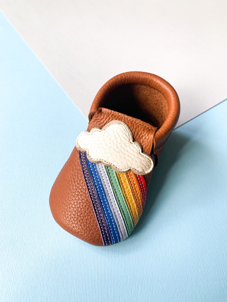 May include: A brown leather baby moccasin with a rainbow and cloud design. The moccasin has a white cloud with a rainbow of colors underneath it.
