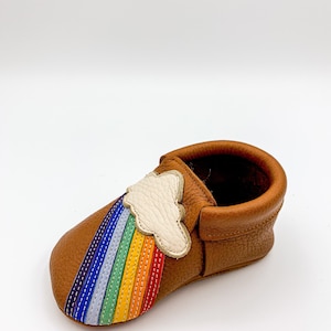 May include: Brown leather baby moccasins with a rainbow and cloud applique. The rainbow is stitched with colorful thread and the cloud is white.