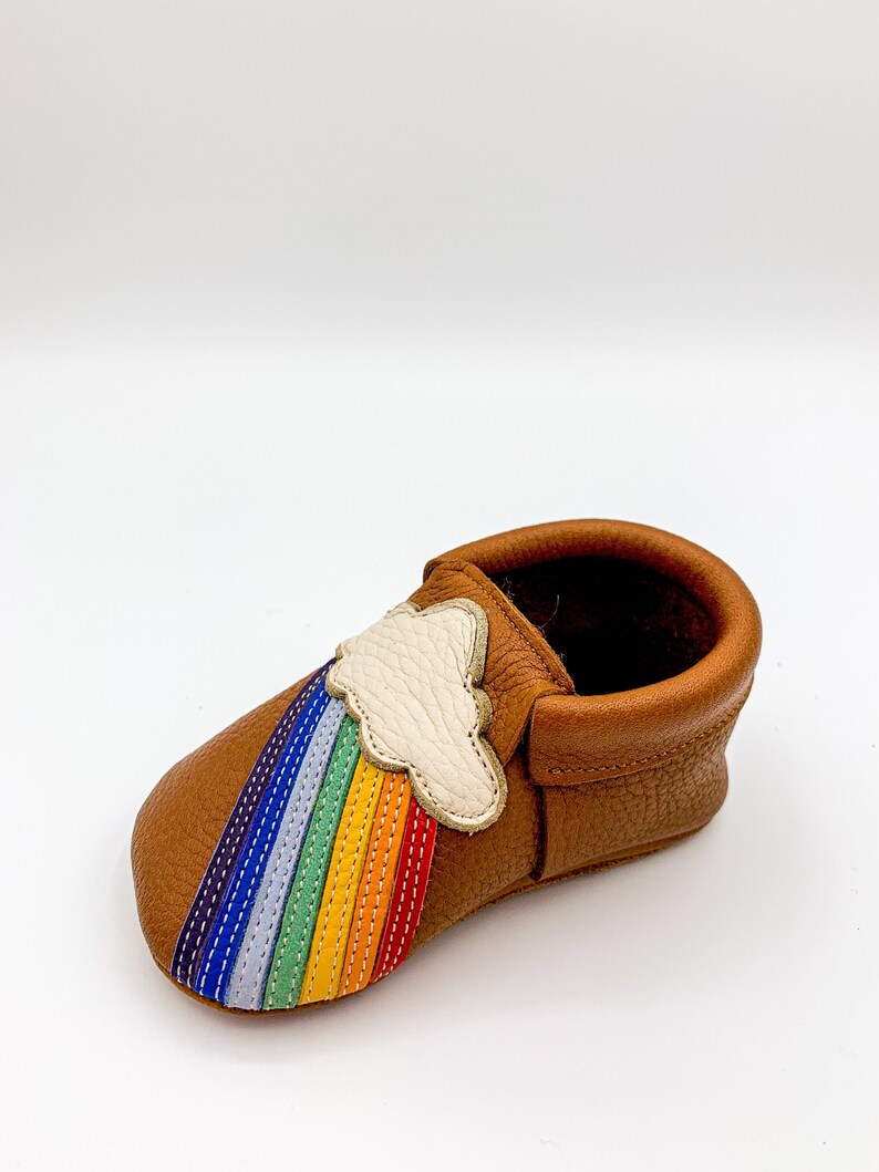 May include: Brown leather baby moccasins with a rainbow and cloud applique. The rainbow is stitched with colorful thread and the cloud is white.