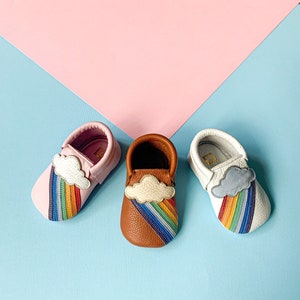 May include: Three pairs of toddler moccasins with a rainbow and cloud design. One pair is pink leather, one is brown leather, and one is white leather.