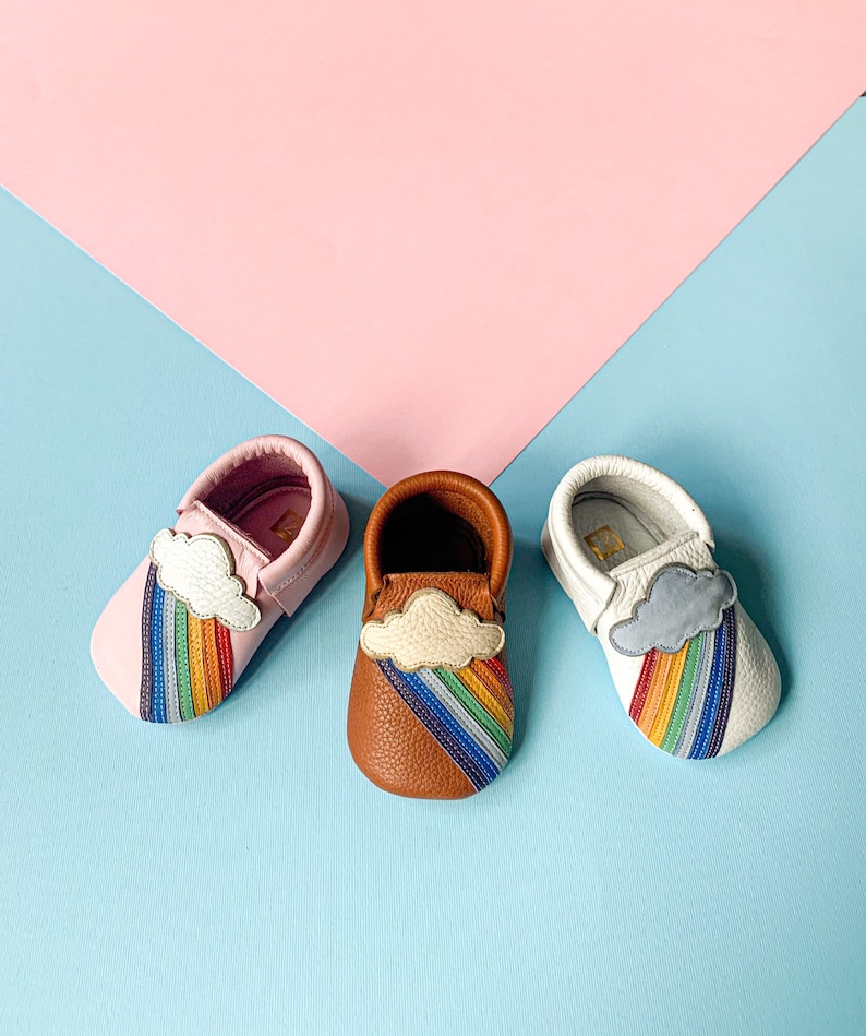 May include: Three pairs of toddler moccasins with a rainbow and cloud design. One pair is pink leather, one is brown leather, and one is white leather.