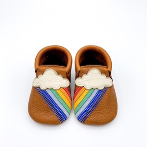 May include: A pair of brown leather baby moccasins with a white cloud and rainbow design. The rainbow is made of colorful stitching.
