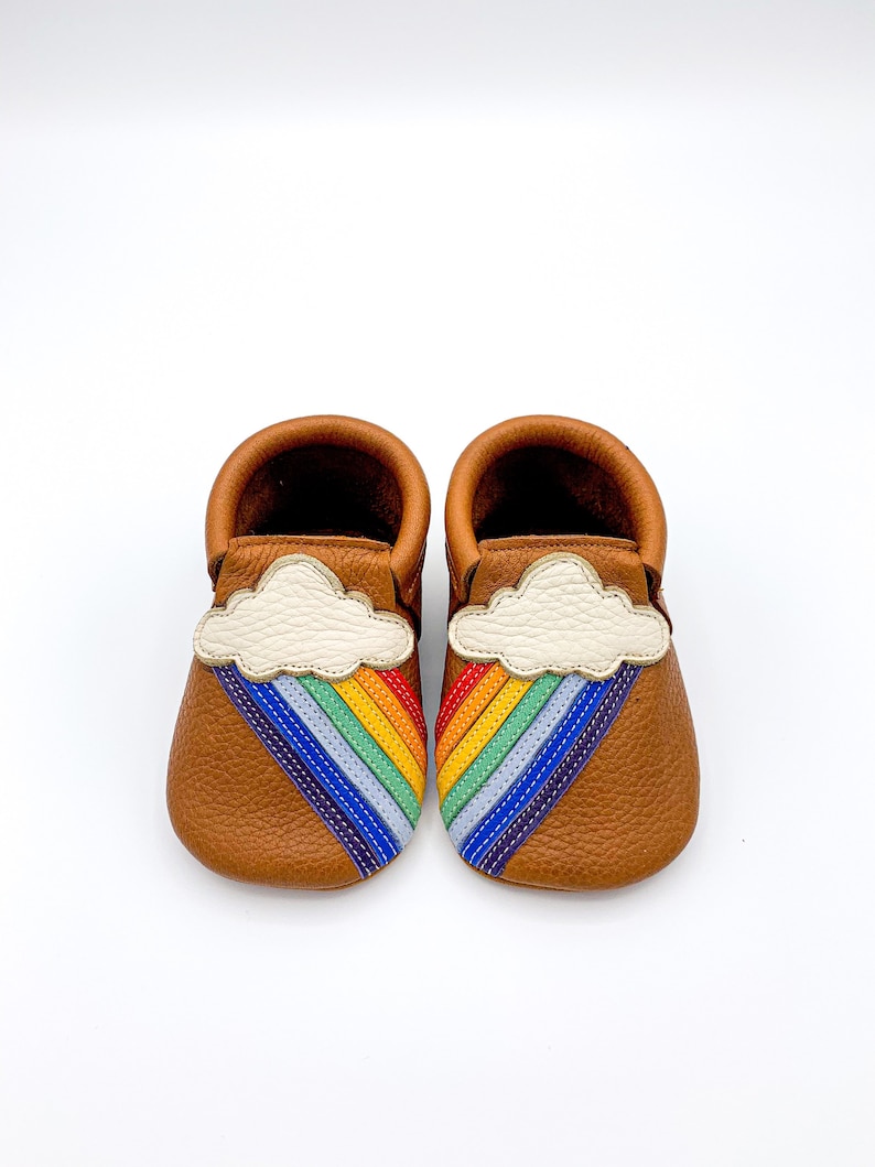 May include: A pair of brown leather baby moccasins with a white cloud and rainbow design. The rainbow is made of colorful stitching.