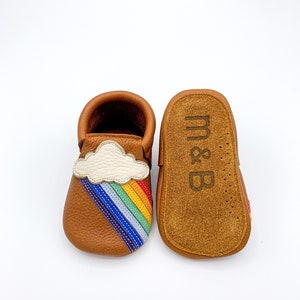 May include: A pair of brown leather baby moccasins with a rainbow and cloud applique. The moccasins are soft and flexible, perfect for little feet.