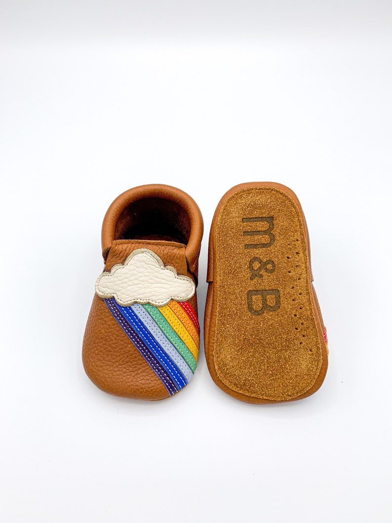 May include: A pair of brown leather baby moccasins with a rainbow and cloud applique. The moccasins are soft and flexible, perfect for little feet.