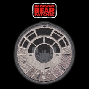 May include: A gray and white embroidered patch depicting the Millennium Falcon cockpit. The patch is circular and has the text "PADAWAN BEAR PATCHES" in red letters.