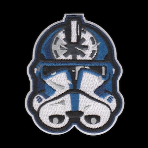 Puede incluir: Parche bordado con un dise&ntilde;o de casco azul y blanco. El parche tiene un borde negro y representa un casco estilizado con detalles intrincados. El dise&ntilde;o incluye un emblema circular y rasgos faciales sim&eacute;tricos, sobre un fondo negro.