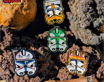 May include: Enamel pins featuring Star Wars clone trooper helmets in various colors, including orange, green, and blue. The pins are displayed on a textured, dark brown surface. The top left corner has a logo that says "Padawan Bear Patches & Pins".