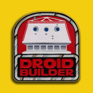 May include: Embroidered patch featuring a white and gray droid design on a red background. The patch has the words "DROID BUILDER" in red block letters. The patch is bordered with a black and gray stitched edge, set against a yellow background.