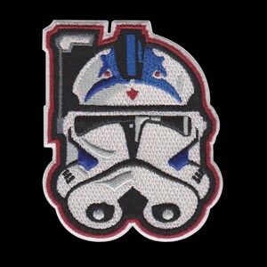 Puede incluir: Parche bordado con un dise&ntilde;o de casco blanco y azul estilizado, con un borde negro y rojo. El parche tiene una apariencia detallada y texturizada, con un fondo negro. El dise&ntilde;o recuerda a un casco de soldado clon.