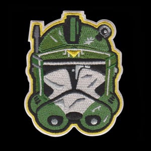 Puede incluir: Parche bordado con un dise&ntilde;o de casco verde y blanco con detalles amarillos y negros. El parche tiene un estilo detallado de dibujos animados, sobre un fondo negro. La forma distintiva del casco sugiere un tema de ciencia ficci&oacute;n o fantas&iacute;a.