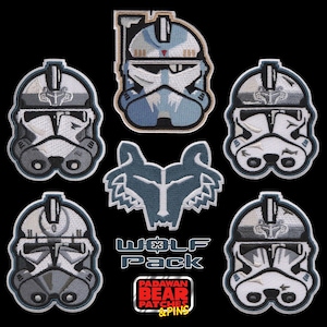 May include: A collection of six embroidered patches featuring five stylized clone trooper helmets in shades of white, gray, and blue, and one wolf head emblem. The text WOLF PACK and PADAWAN BEAR PATCHES & PINS is printed in white and red.