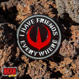 以下が含まれることがあります： 「I HAVE FRIENDS EVERYWHERE」の白い文字が入った、丸い黒と赤のピン。ピンの中央には赤いシンボルがあり、灰色の縁取りがあります。ピンは、質感のある茶色の背景に配置されています。