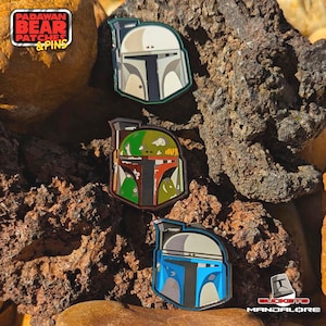 May include: Three enamel pins featuring the helmet design of a popular sci-fi character. The pins are in various colors, including white, green, blue, and red. The pins are displayed on a bed of brown rocks. The top left corner has the text "Padawan Bear Patches & Pins".