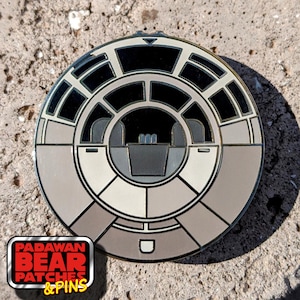 May include: A round, silver-colored pin with a detailed design of the Millennium Falcon's cockpit. The pin is on a light brown, textured surface.