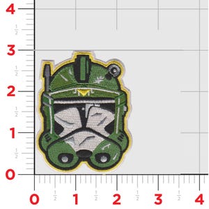 Puede incluir: Parche bordado con un dise&ntilde;o de casco verde y blanco. El parche tiene un borde amarillo y costuras detalladas. El dise&ntilde;o del casco recuerda a un casco de soldado clon. El parche mide aproximadamente 7.6 cm de altura.