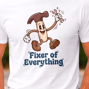 Könnte beinhalten: Weißes T-Shirt mit einer Cartoon-Hammerfigur, die einen Schraubenschlüssel hält. Der Hammer hat einen braunen Kopf und einen beigen Körper, mit braunen Schuhen und weißen Handschuhen. Der Text "Fixer of Everything" ist darunter in Blau gedruckt.