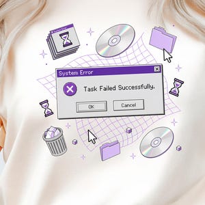Op de afbeelding: Crèmekleurig T-shirt met een retro computer-thema grafisch ontwerp. Het ontwerp bevat een pop-upvenster met de tekst "System Error: Task Failed Successfully", samen met zandlopers, cd's en een prullenbak. De algehele esthetiek is een mix van nostalgie en humor.