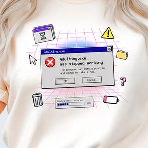 Op de afbeelding: Crèmekleurig T-shirt met een retro computerfoutmelding. De afbeelding toont een pop-upvenster met de tekst "Adulting.exe has stopped working" en diverse computer-thema-iconen. Het shirt heeft korte mouwen en een ronde hals.