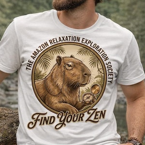 May include: White t-shirt with a circular graphic featuring a capybara, compass, and text that reads "The Amazon Relaxation Exploration Society" and "Find Your Zen". The shirt is worn by a person.