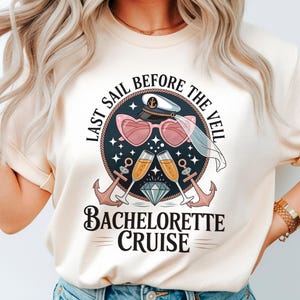Op de afbeelding: Crèmekleurig T-shirt met een grafisch ontwerp voor een vrijgezellencruise. Het ontwerp bevat hartvormige zonnebrillen, champagneglazen, ankers en de tekst "Last Sail Before The Veil" en "Bachelorette Cruise".