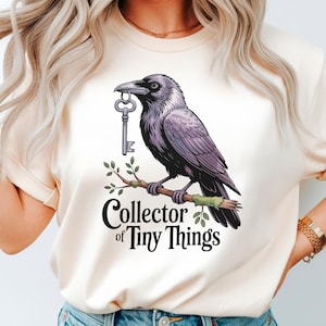 Puede incluir: Camiseta color crema con una ilustración de un cuervo morado sosteniendo una llave en su pico. El cuervo está posado en una rama, con el texto "Collector of Tiny Things" debajo.