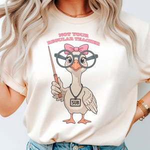 May include: A cream-coloured t-shirt featuring a cartoon goose wearing large round glasses and a pink bow. The goose holds a pointer and wears a lanyard with a tag that reads SUB. Above the goose, the text reads NOT YOUR REGULAR TEACHER.