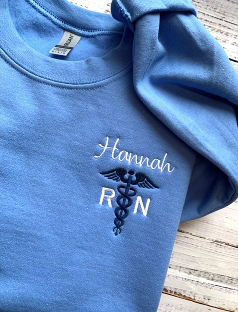 Pu&ograve; includere: Una felpa blu chiaro con il nome "Hannah" e le lettere "RN" ricamate in bianco. Un simbolo di caduceo medico blu &egrave; ricamato tra le lettere.