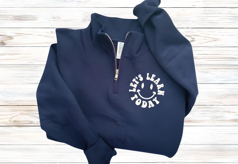May include: Navy blue quarter-zip sweatshirt with a white smiley face graphic and the text "LET'S LEARN TODAY". The sweatshirt has a zipper closure and long sleeves. The background is a light wood grain.