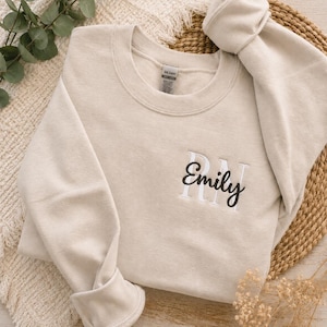 May include: A beige crewneck sweatshirt with the name "Emily" embroidered in black script with white block letters. The sweatshirt is folded on a woven mat with greenery and dried flowers in the background.