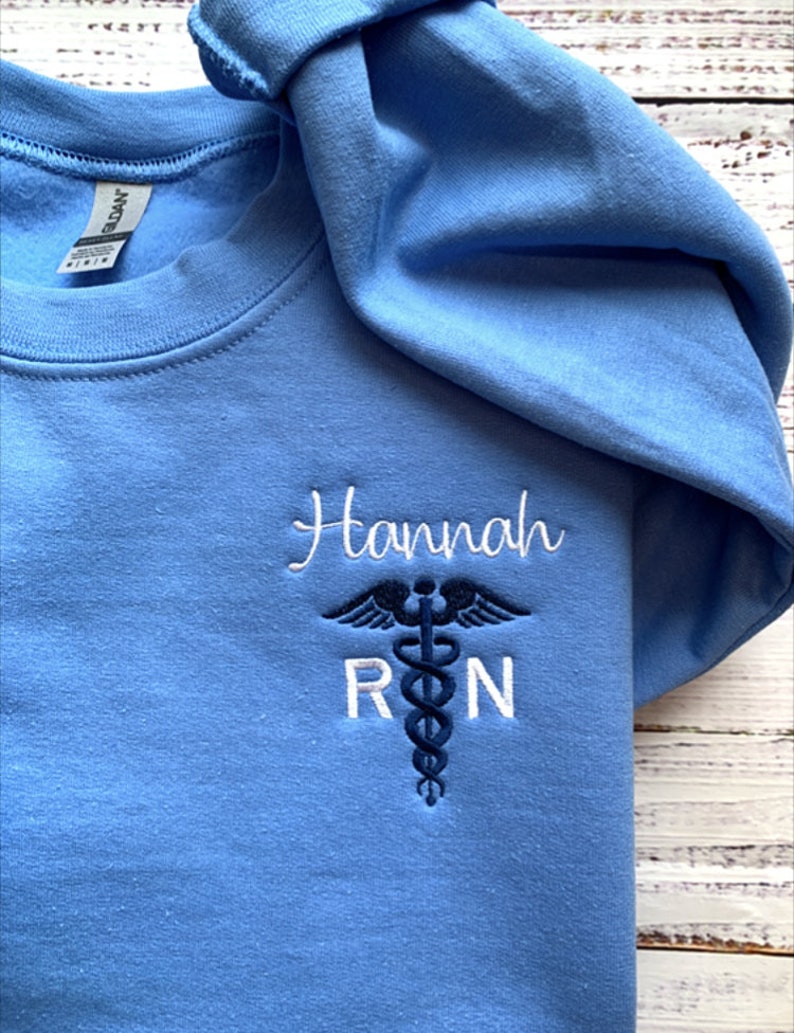 Pu&ograve; includere: Felpa blu chiaro con il nome "Hannah" e le lettere "RN" ricamate in bianco con un simbolo di caduceo medico blu.