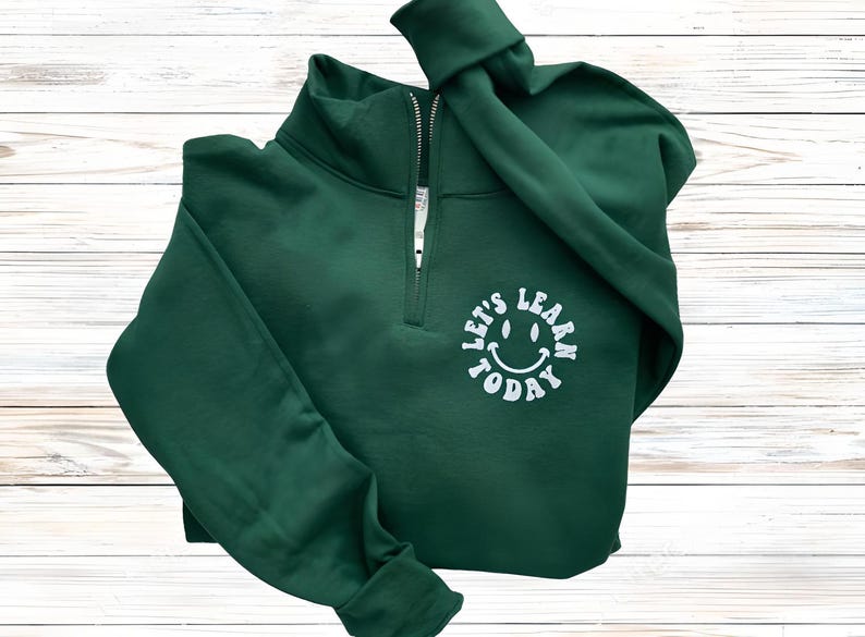 May include: A dark green pullover sweatshirt with a quarter-zip neckline. The sweatshirt features the text "LET'S LEARN TODAY" in a white, circular design with a smiling face. The garment is folded on a light wood-grain surface.