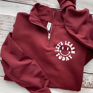 May include: A maroon quarter-zip sweatshirt with the text "Let's Learn Today" in a white, smiling face design. The sweatshirt has a silver zipper and a folded collar. The sleeves are long and folded.