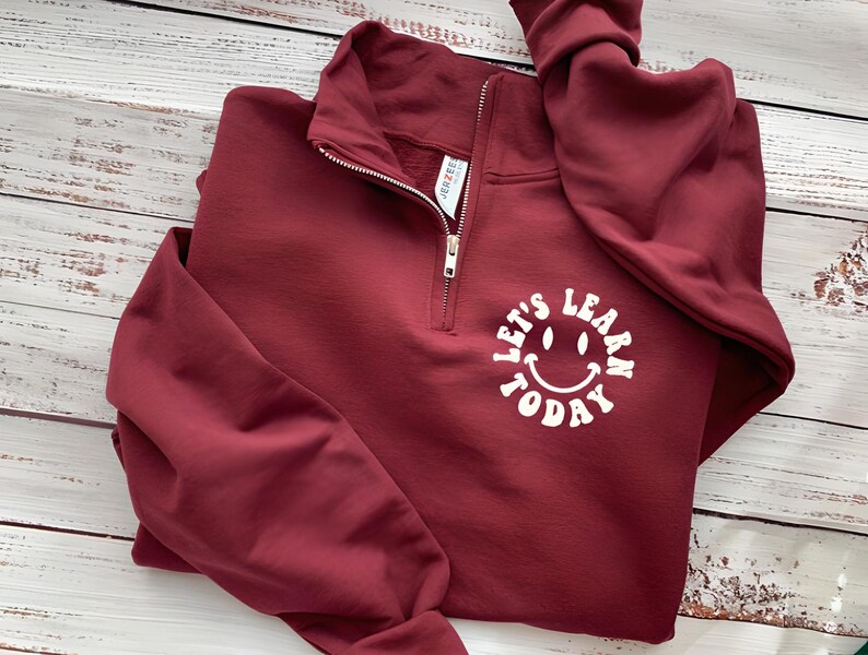 May include: A maroon quarter-zip sweatshirt with the text "Let's Learn Today" in a white, smiling face design. The sweatshirt has a silver zipper and a folded collar. The sleeves are long and folded.