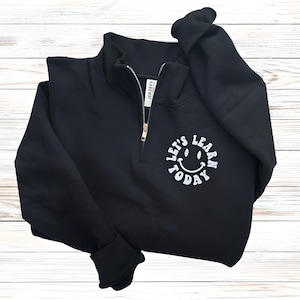 May include: Black quarter-zip sweatshirt with a white smiley face graphic and the text "LET'S LEARN TODAY". The sweatshirt has a zipper closure and ribbed cuffs. The garment is made of a soft material.