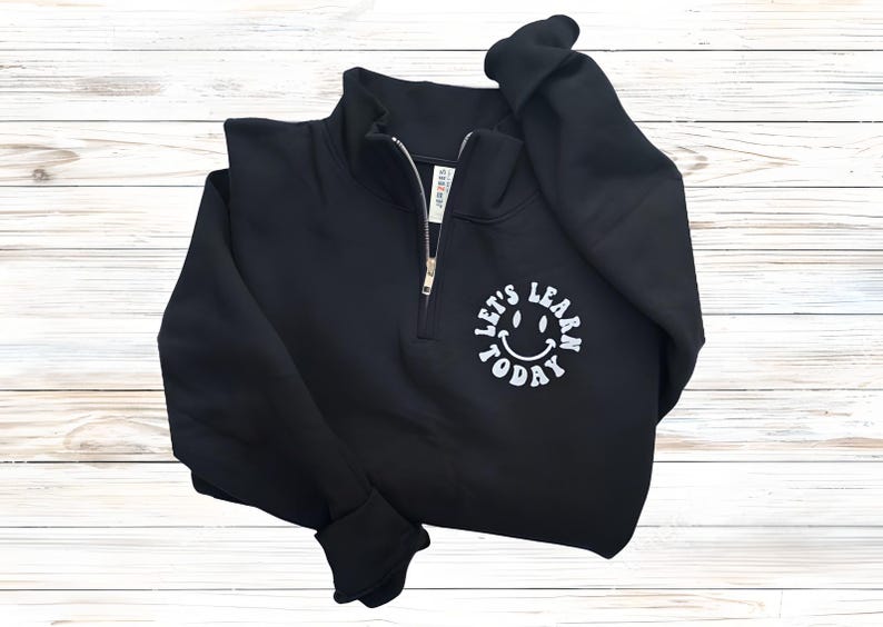 May include: Black quarter-zip sweatshirt with a white smiley face graphic and the text "LET'S LEARN TODAY". The sweatshirt has a zipper closure and ribbed cuffs. The garment is made of a soft material.