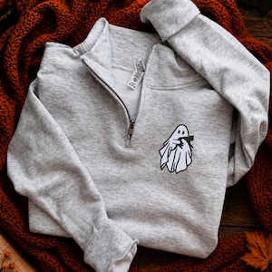 May include: A light gray quarter-zip sweatshirt featuring a small black and white graphic on the left chest. The graphic depicts a white ghost wearing a sheet and holding a small black cat. The sweatshirt has a silver metal zipper at the neck.