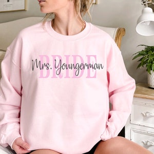 May include: Pink sweatshirt with the text "Mrs. Youngerman" and the word "BRIDE" in pink letters.