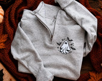 Op de afbeelding: Een lichtgrijs Halloween-sweatshirt met een halslijn met een kwartrits. Het ontwerp toont een witte geest, zwarte vleermuizen en sterren. Het sweatshirt is opgevouwen op een roestkleurige gebreide deken.