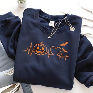 May include: Navy blue sweatshirt with an orange Halloween-themed design. The design includes a jack-o'-lantern, a heart, bats, and a heartbeat line. A silver necklace with a pendant is also visible.