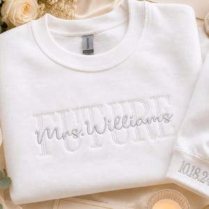 May include: A white crewneck sweatshirt featuring light gray embroidery on the chest that reads FUTURE with the cursive text Mrs. Williams layered over it. The sleeve cuff is embroidered with the date 10.18.24 in matching light gray thread.
