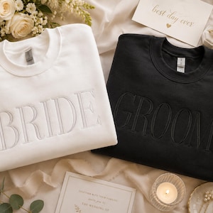 May include: A white crewneck sweatshirt with the word "BRIDE" embroidered in a light gray color. Next to it is a black crewneck sweatshirt with the word "GROOM" embroidered in a matching light gray color. The sweatshirts are on a cream-colored surface.