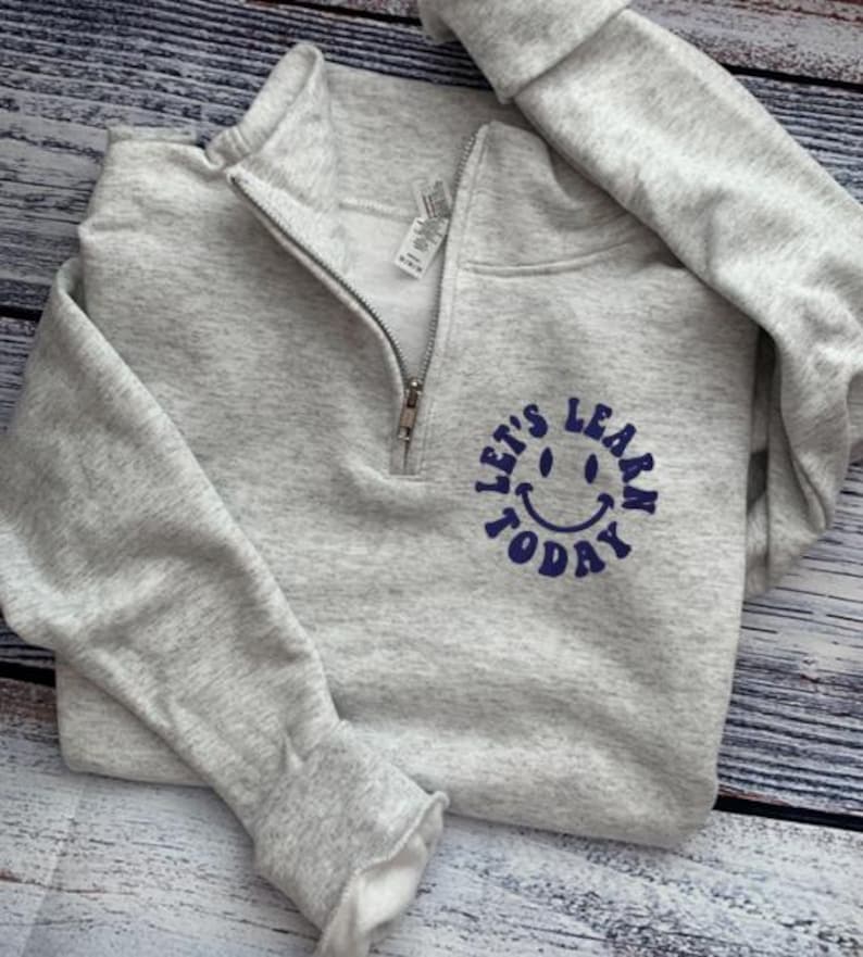 May include: A light gray sweatshirt with a quarter-zip neckline. The sweatshirt features a navy blue graphic of a smiling face with the words "LET'S LEARN TODAY" in a circular design. The sleeves are cuffed.